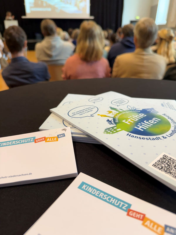 Im vordergrund: Broschüre Frühe Hilfen Lüneburg und Post-it Block Kinderschutz geht alle an; im Hintergrund: viele Teilnehmende schauen sich eine Präsentation auf der Leinwand an